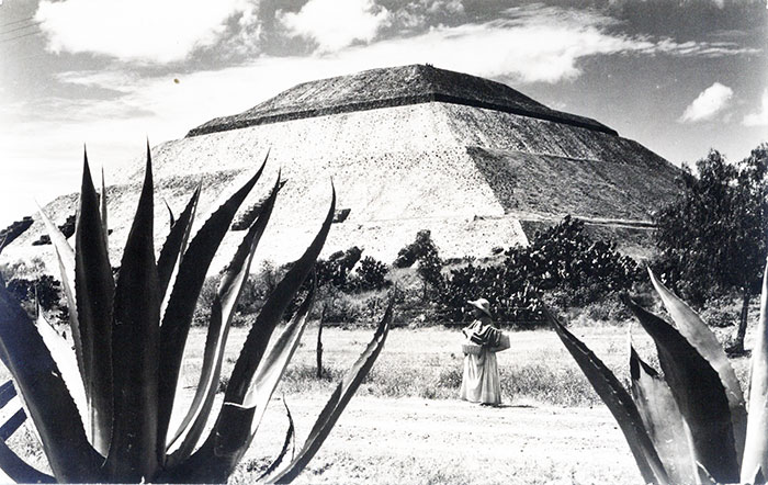 Postcard photo of the Pyramid of the Sun, an indigenous woman with hat, shawl and skirt,
two leaves of agave and shrubs.